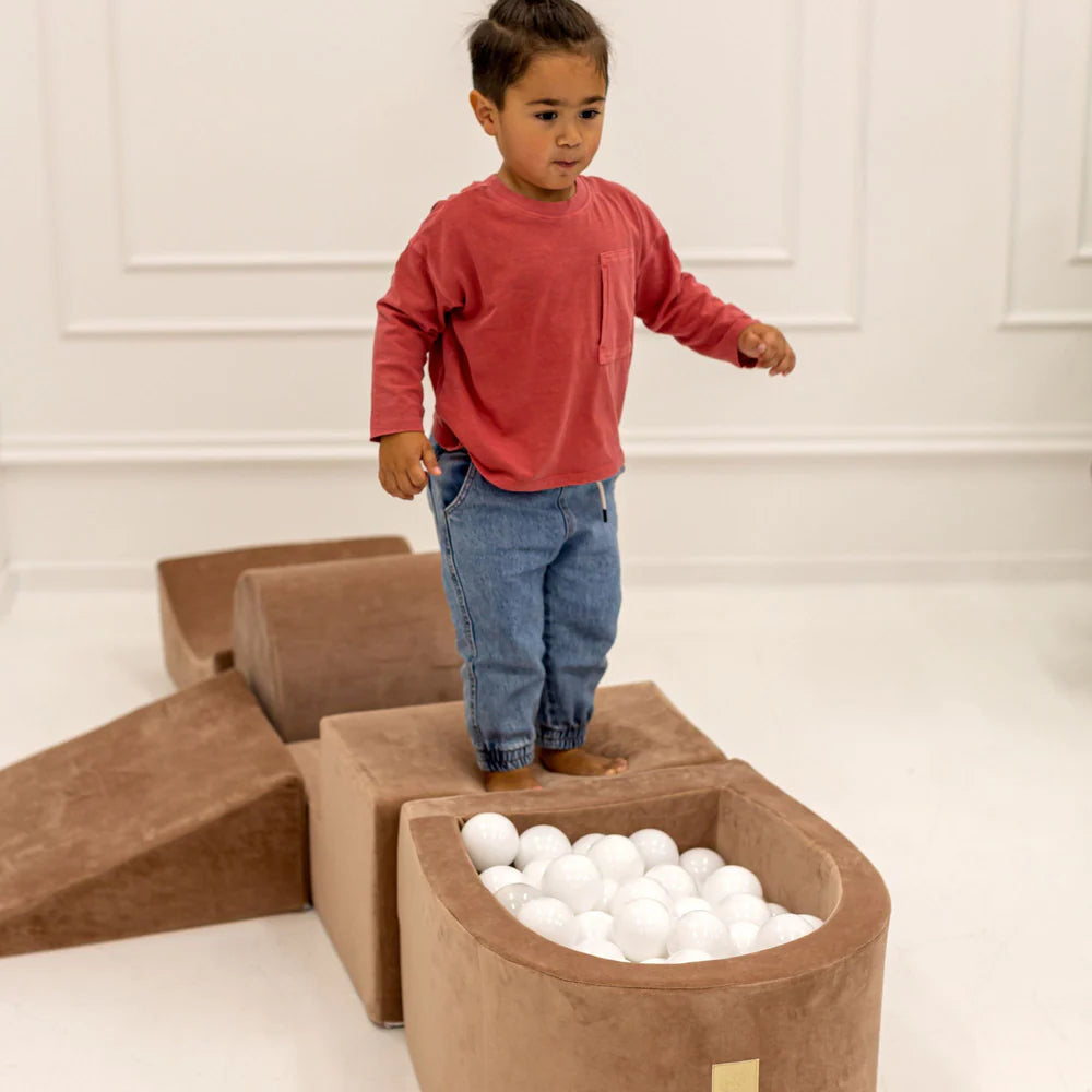 Child playing with a ball pit made of brown foam blocks in a minimalistic room.