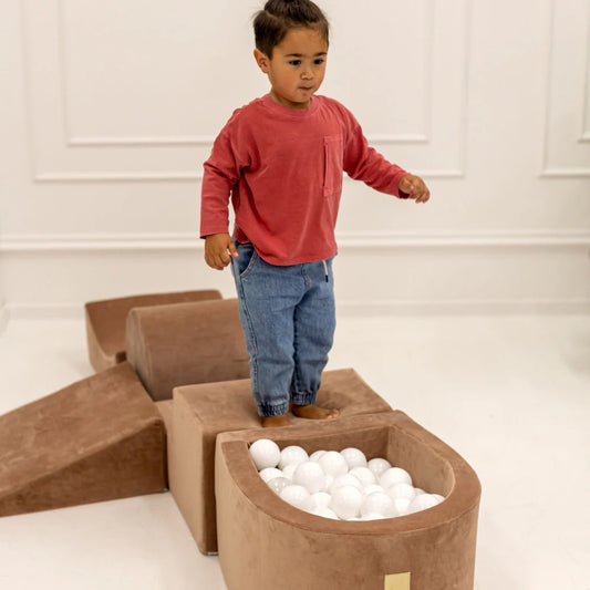 Child playing with a ball pit made of brown foam blocks in a minimalistic room.