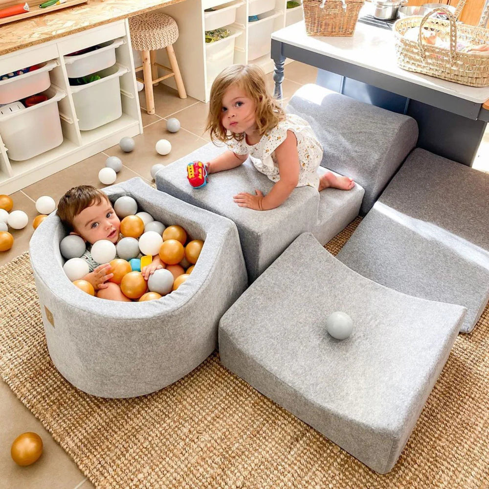 Two children playing with colorful balls in a ball pit surrounded by gray cushioned furniture.