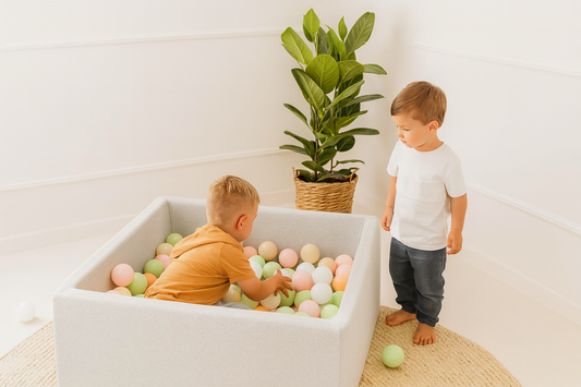 Two children playing with colorful balls in a white container, with a plant in the background.