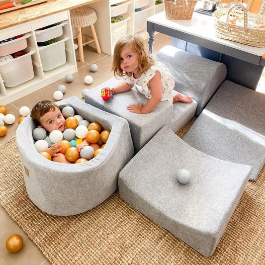 Two children playing with colorful balls in a ball pit surrounded by gray cushioned furniture.